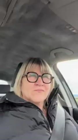 The video depicts a middle-aged woman with blonde hair, wearing glasses and a black puffer jacket, sitting in the driver's seat of a car. She is wearing a seatbelt. She looks directly at the camera with a slight smile and then her expression becomes more serious. The car appears to be in motion. Outside the window, a rural landscape with green fields can be seen. The car's interior is visible, including the ceiling with a textured gray fabric.