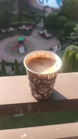A short video captures a cup of coffee placed on a wooden railing. The coffee appears to have a frothy top. In the background, a resort-like setting unfolds with a swimming pool, green trees, and buildings with red-tiled roofs. Several people are visible in the background, some near the pool and others near the buildings. The lighting suggests it is either early morning or late afternoon, with the sun casting a warm glow on the scene. The overall atmosphere is serene and relaxed, evoking a sense of vacation or leisure.
