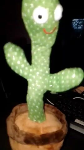 A plush toy cactus with large white eyes and a red smile is positioned in a pot. The cactus has two arms extended outwards and is illuminated by flashing green and pink lights. The background is dark, with a keyboard visible to the right of the toy. The cactus appears to be swaying or dancing.