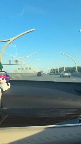 The video shows a view from inside a car driving on a highway on a clear, sunny day.  Several cars are visible ahead in the same direction, and there are distinctive, curved white structures lining the highway. On the dashboard in the foreground is a small, white cat figurine with purple headphones. The dashboard is black leather and has a strip of faux wood trim. The sky is clear and blue.