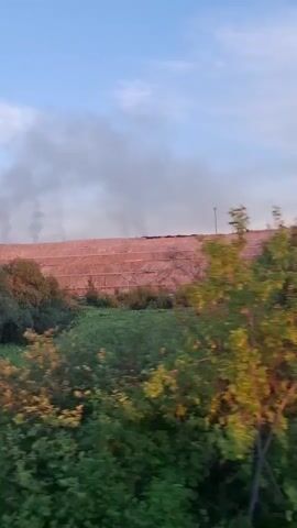 The video shows a landscape scene with a large, terraced embankment in the background, possibly a mining site or waste dump, with a significant plume of dark smoke rising from its crest. The embankment appears to be a reddish-brown color and is covered with some sort of netting or covering material. Green vegetation, including trees and tall grass, grows in front of the embankment and along the edge of a body of water, likely a river or pond, in the foreground. The water reflects the sky and the embankment. The sky is a pale blue with some wispy clouds. The time of day appears to be late afternoon or early evening, given the soft lighting. The overall mood is somewhat somber due to the smoke, which suggests an industrial activity or an uncontrolled event like a fire. There is no visible text in the media. An excavator or similar piece of heavy machinery is faintly visible on the top of the embankment. The scene is set in Achinsk, Russia.