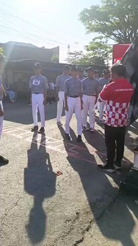 The video shows a group of young males in uniforms standing in a line on an asphalt surface. They appear to be participating in an event. A man in a red shirt with white and red checkered accents is holding a checkered flag with the word "START" visible on it. The flag is positioned in front of the line of young men. The sun is shining brightly, casting long shadows on the ground. The environment appears to be outdoors, possibly a street or an open area, with buildings and trees visible in the background. The young men are wearing similar outfits, consisting of white pants, light-colored shirts with a patterned design, and hats. One of the young men in the line has a bib with the number "30" on it. The man with the flag is facing the line of participants. The overall scene suggests the beginning of a race or competition.