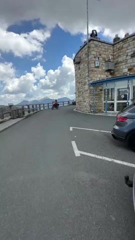 A panoramic video captures a scenic mountain road with a stone building on the right. A motorcycle with two riders is visible on the road ahead. The building features a blue awning and glass doors, with "Ausgang" visible on one door. Several people are gathered near the entrance, some looking at displays inside. A dark-colored SUV is parked on the right side of the road. The background reveals snow-capped mountains under a partly cloudy blue sky.