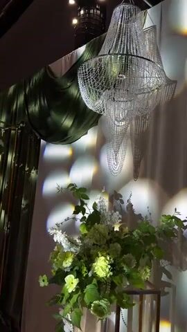 The video shows a dimly lit room with a large, elaborate crystal chandelier hanging from the ceiling. The chandelier is multifaceted and reflects light, casting dappled patterns on the wall. To the left, there is a dark green drape that is partially drawn. In the foreground, there is a large floral arrangement consisting of various shades of green and white flowers and foliage. The flowers are lush and dense, creating a full and luxurious display. The overall atmosphere is elegant and sophisticated, possibly depicting a decorative element for an event or a sophisticated interior.