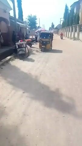 The video shows a street scene in what appears to be a residential area with a dirt road.  A yellow auto rickshaw (tuk-tuk) is parked on the side of the road. There are a few people near a building on the left side of the frame, and a small cart is parked nearby. A motorcycle with two riders passes by on the right side of the road. The area is sunny and the sky is light. There are buildings with fences along the side of the road, and some tall trees are visible.