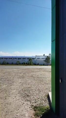The video shows a wide, open, gravel-covered area under a clear blue sky. In the distance, a long, white building with a blue roof is visible, bordered by a white fence and trees. To the right, a tall, green metal structure is partially visible in the foreground. The ground in the immediate foreground is paved and has several puddles of water. There are some bags and containers, possibly waste or materials, near the paved area. In the far distance, there are utility poles and possibly mountains. The overall scene appears to be an industrial or semi-urban outdoor space on a sunny day.