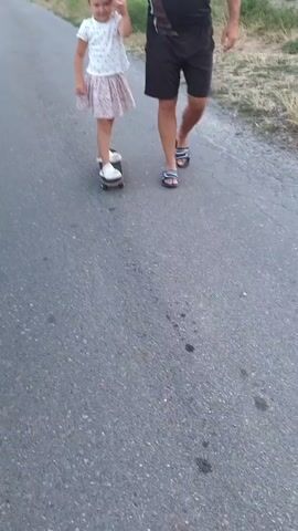 A young girl on a skateboard is being helped by an adult. She is holding the adult's hand while balancing on the skateboard. The adult is wearing shorts and sandals. The girl is wearing a white t-shirt and a floral skirt. They are on a paved path with grass and bushes on the side.