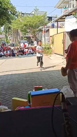 A young boy in a white t-shirt and black pants is performing a dance routine in the middle of a street. He is facing a group of people sitting on the ground to his left. Another person, wearing a red shirt and light-colored pants, stands to the boy's right, holding a yellow object. In the foreground, a stage or platform is visible with colorful boxes and a microphone. The background shows buildings and trees under a clear sky. The scene appears to be an outdoor event.