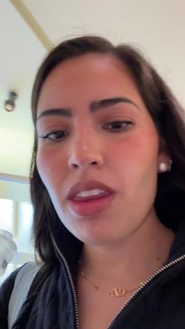 A young woman with long brown hair, fair skin, and noticeable blush is holding and drinking from a clear plastic cup with a tan-colored liquid inside. The cup has a label with the name "Valeria" written on it. She is wearing a black zip-up hoodie, a gold necklace with a pendant, and pearl earrings. The camera is held at a low angle, looking up at her face. The background shows a ceiling with air conditioning vents and light fixtures. In some frames, a blurred figure of another person is visible in the distance. The woman takes a sip of the drink and looks directly at the camera.