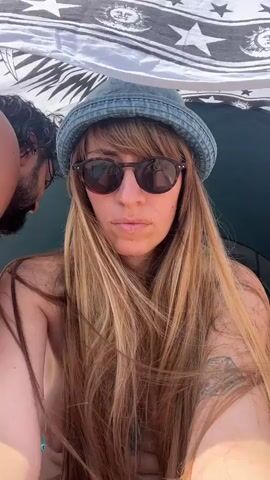 The video shows a close-up of a woman with long blonde hair and sunglasses, wearing a bucket hat. She is under a canopy with a patterned fabric. A person's shoulder and ear are visible on the left side of the frame. The background is a bright, hazy sky, suggesting an outdoor, sunny environment, likely a beach or similar location. The woman appears to be looking directly at the camera.
