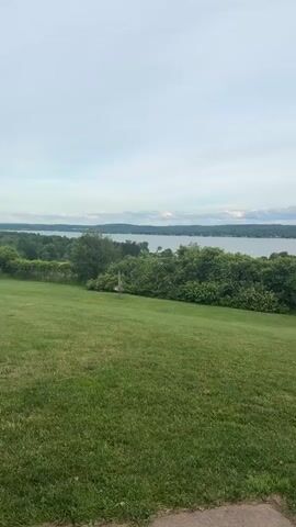 The video shows a panoramic view of a grassy hill overlooking a large body of water. The sky is overcast with scattered clouds. There is a dense line of green trees and bushes separating the grassy area from the water. In the foreground, the grass is neatly mowed. There is a small red and white object that appears to be a bird feeder on a pole in the middle of the frame. In the distance, the water stretches to the horizon, with a faint outline of hills on the far side. The overall impression is one of a serene, natural landscape.