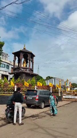 A woman wearing a blue and green sari walks past a dark-colored SUV. Behind the SUV, a dark-colored decorative structure with a golden statue is visible. Yellow flags are displayed along a black fence. Further in the background, a building with several golden statues on a raised platform is seen, with a tall tower in the distance. A police barricade with Marathi text is in the foreground, and several people are on motorcycles and a car. The sky is cloudy.