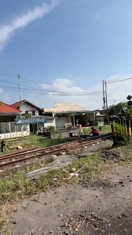 The video shows a railway track with a small building next to it. The building has a sign that reads "NANGGULANGIN" in Indonesian. There are also some yellow and black striped poles near the track, which might indicate a railway crossing. The environment appears to be a rural or suburban area with some vegetation and houses in the background. The sky is blue with some clouds. There are no people visible in the video.