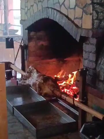 A whole animal carcass, likely a pig, is being spit-roasted over an open fire within a brick fireplace. The carcass is skewered on a long wooden rod that is supported by metal stands on either side, allowing it to rotate. The fire beneath the carcass is vibrant with flames and glowing embers. Below the roasting carcass, two rectangular metal trays are positioned to catch any drippings. The fireplace is built with rough-hewn stone and brick. To the left of the fireplace, a black mechanical device with a rotating arm is visible, likely part of the spit-roasting mechanism. The environment appears to be indoors, possibly a restaurant or a commercial kitchen, with a window and some other background elements suggesting this. The video captures the slow, steady rotation of the carcass as it cooks.