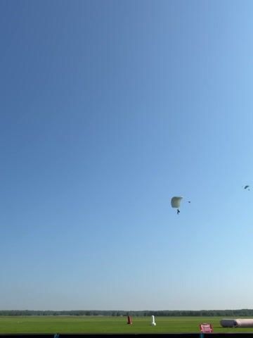 A wide shot shows a clear blue sky over a green field. Two parachutists are descending. The closest parachutist is in the middle left of the frame, with a white parachute and a person hanging below. Further to the right, a second, smaller parachute is visible. The ground below is a grassy field with some scattered objects, including red and white markers, and a sign on the right side of the frame. The horizon is lined with trees.