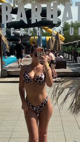 The video shows a woman taking a selfie in front of a mirror at what appears to be a beach club or water park. She is wearing a brown and white patterned bikini. In the reflection of the mirror, a sign that reads "BARHAT BEACH CLUB" is visible. Behind her, there are water slides, people in swimwear, and palm trees. The woman is posing for the camera, holding her phone to take the picture.