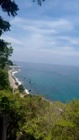 A high-angle, wide shot shows a coastal road alongside a bright blue ocean under a cloudy sky. Lush green trees in the foreground frame the scene. The road, which curves along the coastline, has very few cars on it. Waves crash against a rocky shore interspersed with patches of sand and greenery. Several large rocks are visible in the water close to the shore. In the distance, buildings can be seen along the coastline. The overall impression is a beautiful, somewhat remote coastal landscape.