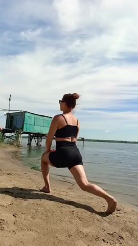 A woman with auburn hair pulled into a bun and wearing sunglasses, a black sports bra, and black shorts is captured from behind on a sandy beach. She is performing a lunge exercise with her right leg forward and bent, and her left leg extended back. Her arms are bent at the elbows. The background features a calm body of water, a bright blue sky with scattered white clouds, and a small green shack built on stilts over the water, with a wooden pier extending from it. Another person, a man in swim trunks, is visible walking near the water's edge in the distance. The scene appears to be set on a sunny day, likely in the late morning or early afternoon, given the bright lighting. The location is identified as Vyazovka, Russia. The overall mood is one of active recreation and enjoying the outdoors. There is no visible text in the media file.