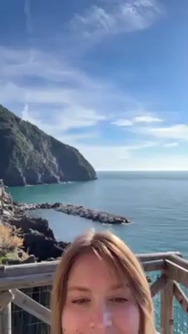A fair-skinned woman with long blonde hair stands smiling on a wooden deck or balcony, overlooking a rocky coastline and the sea. She is wearing a pink top and a dark jacket. Behind her is a picturesque view of a rocky cliff covered with green vegetation, and the calm, blue ocean under a sunny, partially cloudy sky. The railing of the deck is made of light-colored wood.