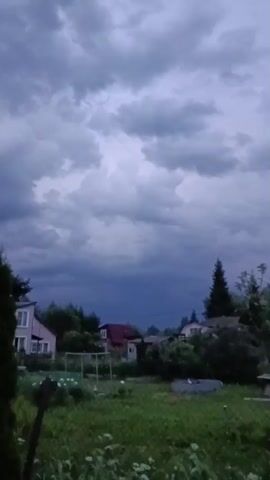 A video shows a cloudy sky over a yard with houses and trees. The clouds are dark and appear to be heavy with rain. The yard is green and overgrown with grass and weeds. There are a few houses and trees in the background. The video is short and does not show any people or animals. The lighting is dim, suggesting it is either dusk or a stormy day.