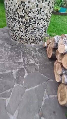 The video begins with a blurry shot of chopped logs stacked on a stone patio, with grass visible in the background. The camera then pans to the right, focusing on a circular mosaic-covered base with a wooden lid that has two metal handles. The lid appears weathered and aged, with some peeling paint. The background shows a grassy area with some greenery and faint glimpses of outdoor equipment. The camera continues to move, providing closer views of the wooden lid and the mosaic base. The video ends with a close-up shot of the wooden lid.