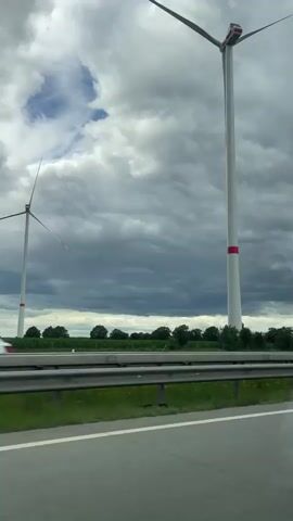 A video shot from a moving vehicle shows a view of a road with a metal guardrail on the right side. Beyond the guardrail, there is a grassy area and a line of trees. In the background, two large wind turbines are visible against a cloudy sky. The closest wind turbine is tall and white with red bands around its base. Its blades are angled upwards. A white car is driving on the road in the same direction as the vehicle from which the video is being recorded. The sky is filled with dark, heavy clouds with some lighter patches. The overall lighting suggests an overcast day.