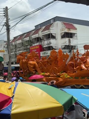 The video shows a large, orange, intricately carved float that appears to be made of wax. The float is decorated with figures of people and animals, possibly depicting a mythological or religious scene. It is on display outdoors, possibly during a festival or parade, with several colorful umbrellas in the foreground providing shade. In the background, there are buildings with awnings and electrical wires crisscrossing overhead. A crowd of people, some holding umbrellas, are visible gathered around the float. The overall atmosphere suggests a public event taking place during the day.