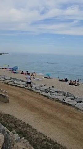 The video shows a beach with a number of people sunbathing, swimming, and relaxing. The beach is sandy and there are several large rocks separating the sand from a dirt path. A man wearing a white shirt and black shorts stands on the rocks with a small dog next to him. Further out in the ocean, there are boats sailing and a few distant objects that could be more boats or buoys. The sky is blue with some scattered white clouds.