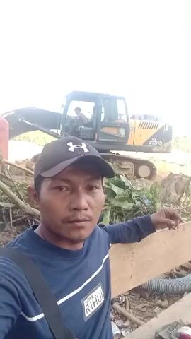A man wearing a blue long-sleeved shirt and a black baseball cap is in the foreground, looking directly at the camera. He has a dark complexion and short dark hair. A black strap is visible across his chest. Behind him, an excavator with a yellow body and black tracks is positioned on a dirt mound. A person is visible inside the excavator's cabin. The environment appears to be outdoors with vegetation and debris scattered around. A red cylindrical object is partially visible to the left of the excavator.
