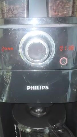 A close-up, slightly blurry video shows the control panel of a black Philips coffee maker. The digital display shows "2000" on the left and "01:38" on the right. A silver dial is in the center, and a small circular button is below the right-hand display. The coffee maker has two compartments for coffee beans at the top. The camera briefly pans out, showing the coffee maker is on a countertop, with other objects visible in the background. The overall lighting is dim.