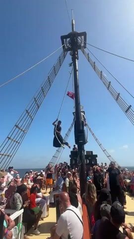A person in dark clothing swings on a rope attached to the mast of a large ship. The person is suspended in mid-air, upside down, with their legs bent. They are on a pirate ship, with many people on deck watching the performance. The sky is blue and clear, and the ocean is visible in the background. The rope appears to be attached to a rigging system of the mast. The person is actively engaged in a dynamic movement as part of an apparent show or entertainment. The crowd is gathered around the base of the mast, looking up at the performer.