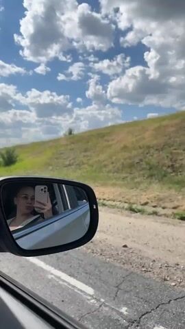 The video shows a view from inside a moving car. The camera is focused on the passenger-side mirror, which reflects a young woman taking a selfie with her phone. She is wearing glasses and a light-colored top. Outside the car, a grassy hill under a cloudy blue sky is visible. The car is on a dirt road next to a paved road.