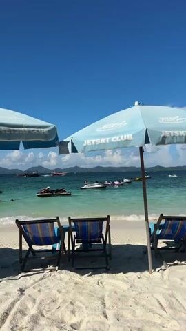 The video shows a tropical beach with white sand and clear blue water. Three beach chairs are set up on the sand, facing the ocean. Two light blue beach umbrellas provide shade. In the distance, several jet skis are visible on the water, along with some boats. The sky is a clear, bright blue with a few white clouds. The overall impression is one of a sunny, relaxing day at the beach.