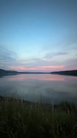 A still shot of a body of water at dusk. The sky is a gradient of deep blue at the top, transitioning to pink and orange hues near the horizon. Wispy clouds are visible. The water's surface is calm, reflecting the colors of the sky. In the foreground, tall dry grass is visible. The scene appears to be a natural landscape with hills or landmasses visible on the distant horizon. There are no people or objects visible in the video. The video is static, with no movement or action.