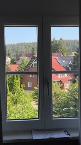 The video pans across a window with a view of a suburban area. The window has a white frame and two panes on each side. A grey curtain is visible on the left side of the window and moves to cover the right side of the window. In the foreground, on the windowsill, a potted plant is visible. Outside the window, there are houses with red roofs, trees, and a forested hillside in the background. The sky is clear and blue.