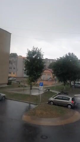 The video shows a rainy day in what appears to be a residential area in Smarhon, Belarus. The scene is an outdoor view, likely from a window, overlooking a street and a small park or courtyard area.

Several objects are visible: cars are parked along the street and in a designated parking area. One silver car is positioned on a grassy traffic island. There are trees, a playground set with swings, and benches in the courtyard. Buildings, likely apartment complexes, line the street. One building is a light brown color, and another is a pale yellow, with multiple windows and balconies. A blue car is parked on the right side of the street.

The primary activity is the heavy rainfall, which is evident by the streaks of water falling and the wet appearance of the pavement and cars. The sky is overcast and grey, indicating the time of day is likely afternoon or late morning, with poor lighting due to the weather. The overall mood is somber and damp, typical of a rainy day. There is a blue and white sign with a "P" symbol, indicating a parking zone, and a smaller sign below it with the number "7".