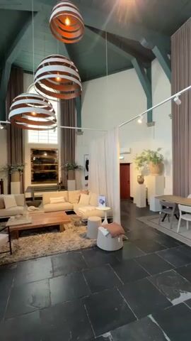 The video showcases the interior of a spacious, high-ceilinged building in Noordwijk, Netherlands, likely a repurposed church or hall, now serving as a furniture and home decor showroom. The room features a dark green vaulted ceiling with exposed beams, white walls, and a dark stone or large tile floor.

Various furniture arrangements are displayed, including a living room setup with light-colored sofas, armchairs, a wooden coffee table, and a shaggy rug. Another area features a dining table with chairs, and decorative elements like poufs and side tables are scattered throughout. The space is adorned with large, distinctive pendant lights made of wooden slats, track spotlights, potted plants, floral arrangements in vases, and framed artwork on the walls. Light-colored fabric screens and a tall brown panel act as room dividers, creating distinct display zones. A wooden pulpit-like structure is visible in the background, hinting at the building's original use.

No people are visible in the video. The lighting is a mix of warm artificial lights and natural light coming from large windows. The overall aesthetic is modern, comfortable, and uses a palette of natural tones such as beige, brown, and white. At the beginning of the video, a partially visible sign reads "ART EXPO" with dates "14 MEI - 7 JUN".