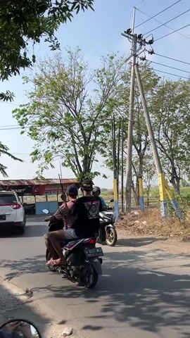 The video shows a street scene with three people on two motorcycles. In the foreground, two individuals are riding a motorcycle, with the person in the back holding a long, thin object, possibly a stick or pole. They are on a paved road. To their right and slightly behind, another motorcycle is visible with a single rider. In the background, there are trees, utility poles, and a structure that appears to be a roadside stall or building. A white SUV is parked on the left side of the road. The environment appears to be an outdoor, semi-rural setting with dry vegetation. The sky is clear and the lighting suggests it is daytime.
