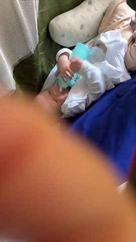 A baby wearing a white onesie and a white hooded garment is lying on their back. The baby is holding a light blue bottle with their right hand and feeding from it with their mouth. A blue blanket is visible beneath the baby. A tan and beige patterned pillow is behind the baby's head, and a portion of a green tent or playpen is visible around the baby. The camera is positioned above and to the side of the baby.