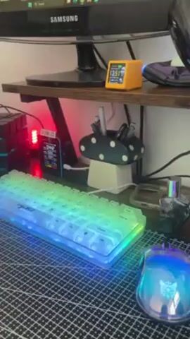 A brightly lit desk setup features a Samsung monitor atop a rustic wooden shelf. A small, square yellow digital clock sits next to a black computer mouse. Below, a white, compact keyboard with rainbow-colored backlighting rests on a checkered mat beside a blue and pink illuminated mouse. A mushroom-shaped pen holder filled with pens and writing utensils are to the right of the keyboard. An electronic device with a red light glowing behind it sits to the left. Electrical cords snake around the various objects.