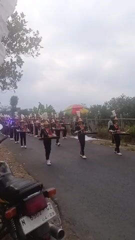 A marching band consisting of approximately twenty to thirty individuals, all wearing matching black and orange uniforms with white hats and footwear, marches down a paved road. Each band member carries a percussion instrument, likely a snare drum, in front of them, holding it with two hands. The band is moving from the left side of the frame towards the right. In the foreground, a portion of a motorcycle is visible on the left side of the road. The environment appears to be rural, with trees and vegetation lining the road. The sky is overcast, with grey clouds dominating the upper portion of the frame. A colorful, striped umbrella or canopy is visible in the background, behind the marching band, and a white banner or flag is partially visible at the top left of the frame. The band members are in formation, with several rows of individuals marching in unison. The overall scene depicts a parade or a marching band performance.