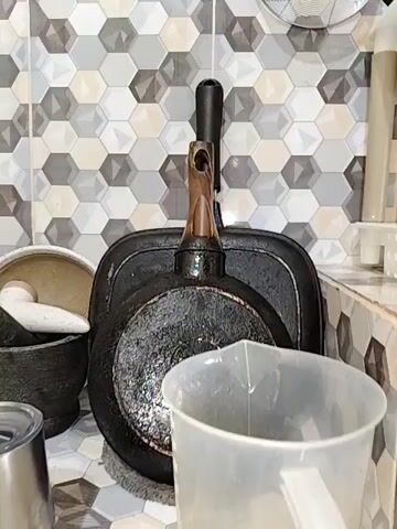 THE VIDEO SHOWS A CLOSE-UP VIEW OF A KITCHEN SETTING. IN THE FOREGROUND, A TRANSPARENT PLASTIC MEASURING CUP IS VISIBLE. BEHIND IT, TWO BLACK FRYING PANS ARE HUNG ON A WALL. THE PANS ARE STACKED VERTICALLY, WITH THE LARGER PAN BEHIND THE SMALLER ONE. THE SMALLER PAN HAS A WOODEN AND METAL HANDLE. THE WALL BEHIND THE PANS IS COVERED WITH HEXAGONAL TILES IN SHADES OF GREY, WHITE, AND BLACK. TO THE LEFT OF THE PANS, A BLACK MORTAR AND PESTLE ARE PARTIALLY VISIBLE.
