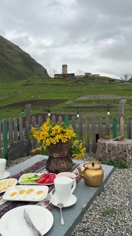 A video shows a table set for breakfast with a scenic mountain view. The table is covered with a patterned rug and features plates of food including sliced cheese, sliced cucumbers and tomatoes, fried eggs, and a bowl of jam. A golden teapot and two white teacups are also on the table, along with a vase of yellow flowers. In the background, a wooden fence encloses a green, grassy field with sheep. Beyond the field, there are several small buildings and a stone tower on a hillside, with snow-capped mountains in the distance. The sky is overcast. The camera pans slowly across the scene.
