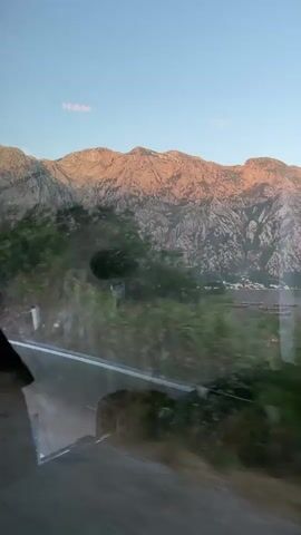 The video shows a view from inside a vehicle, looking out the window at a mountainous landscape at sunset. The sun's rays cast a warm, golden glow on the rugged peaks, highlighting their texture and contours. Lush green trees and foliage line the roadside, blurring slightly due to the motion of the vehicle. A road is visible in the foreground, with a white line marking its edge. The sky is a clear, pale blue with a hint of pink near the horizon. Reflections of the vehicle's interior, including text that appears to read "MAN" and "75", are visible on the glass. A triangular road sign is also partially visible. The overall impression is one of a scenic drive through a natural environment bathed in the soft light of twilight.