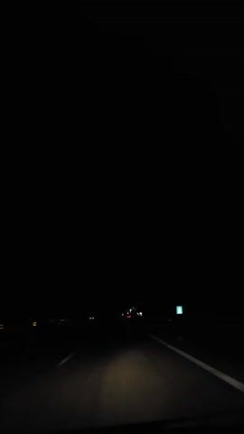A low-angle shot from the driver's perspective shows a car traveling on a highway at night, facing forward. The only visible elements are the road ahead, illuminated by the car's headlights, and the occasional distant headlights of other vehicles. The road has white double solid lines and white dashed lines. The sky above is entirely dark, with no stars or moon visible. The environment appears to be an open, unpopulated area, with no structures or notable landmarks in sight. A brief flash of lightning reveals a cloudy sky over the landscape in the distance.