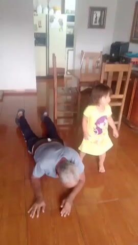 A man is on the floor in a push-up position, while a young girl, wearing a yellow dress with purple accents, sits on his back. The man appears to be an elder, with grey hair and wearing a blue t-shirt and black pants. The girl is young, with dark hair, and appears to be a toddler. The room has tiled flooring and wooden furniture, including chairs and a table, suggesting a home environment. In the background, a refrigerator and cabinets are visible, as well as a framed picture on the wall. The scene takes place indoors, with natural light suggesting daytime. The overall impression is one of playful interaction between the man and the child. There is no visible text in the video.