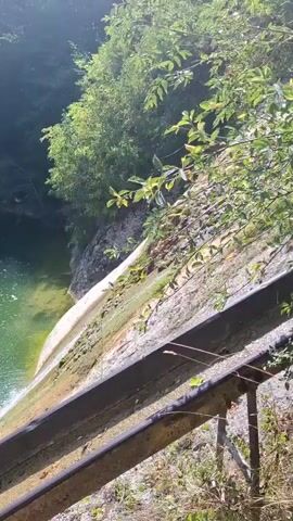A narrow shot pans across a body of water from the edge of a slope. The water is green and appears shallow, with some plant life visible beneath the surface. A waterfall is partially visible on the right side of the frame, where water cascades down a moss-covered concrete structure. The slope itself is covered in vegetation, and a rusty metal railing runs along the bottom of the frame. In the background, trees and rocks suggest a natural, forested environment. The sunlight is bright, casting shadows and highlighting the ripples on the water.