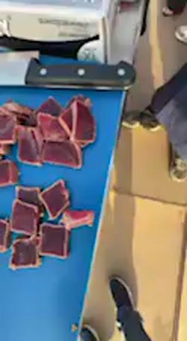 The video captures a close-up of a food preparation and serving station, likely at an outdoor event in Barbate, Spain. A person, whose hand is visible wearing a black glove, is actively placing individual slices of dark-colored, cooked meat onto small white paper plates. These plates, each holding several slices, are then arranged on a light-colored table surface, suggesting they are being prepared for tasting or serving.

In the foreground, a blue cutting board holds a pile of the same sliced meat, alongside a large knife with a black handle. Other objects on the table include a metal container, possibly holding a sauce or liquid, and a white squeeze bottle. In the blurred background, a crowd of people can be seen, indicating a public gathering or festival. A box with the text "KORNNY" is also visible. The scene suggests a lively culinary event, possibly focusing on local seafood, with samples being distributed to attendees. The time of day appears to be bright and clear.