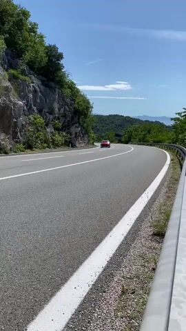 A stationary shot captures a two-lane asphalt road with white solid lines on both sides. On the left, a steep, rocky, tree-covered mountain slopes upward. A white guardrail lines the right side of the road. In the distance, a winding road disappears into a valley of hills and mountains under a clear blue sky. A red car drives past the camera on the left, followed by a white van on the right.