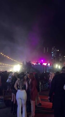 The video shows a crowd of people at a nighttime outdoor event, seemingly a concert or festival. In the center of the frame, a raised platform serves as a stage, with figures silhouetted against bright lights, presumably performers or DJs. The stage is flanked by light towers and illuminated by a purple-hued light that fills the upper portion of the frame. String lights are visible in the background, adding to the festive atmosphere. The crowd, which appears mostly female, is dressed in casual, trendy clothing, with several individuals wearing cowboy hats. People are walking, talking, and observing the performance on stage. The camera remains mostly static, capturing the energy and excitement of the event.